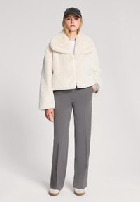 Giacca in pelliccia sintetica color crema con ampio colletto, chiusura a bottone e maniche lunghe, abbinata a pantaloni grigi e sneakers bianche.