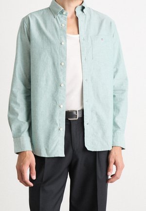 Mężczyzna ubrany w jasnozieloną koszulę zapinaną na guziki na białą podkoszulkę, czarny pasek i czarne spodnie, stojący na jednolitym tle.