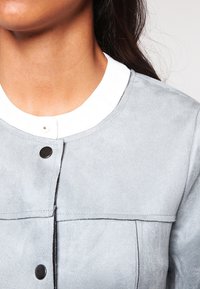 Chaqueta de ante gris claro con un cuello blanco, que cuenta con un cierre de botones a presión y una textura suave. Detalles de costura notables en el pecho.