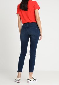 Jeans ajustados de color azul oscuro con bolsillos traseros, combinados con una blusa roja de manga corta y zapatillas tipo mocasín de cuadros, vistos desde atrás.