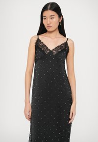 Fekete pántos ruha, finom csipkével díszített nyakkivágással és szórt ezüst díszítésekkel. Az anyag sima tapintású és enyhén fényes.