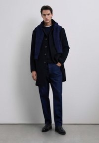 Abrigo negro con un diseño estructurado, usado sobre un suéter azul marino. Una bufanda de punto azul marino cae alrededor del cuello, combinada con pantalones de mezclilla oscuros.