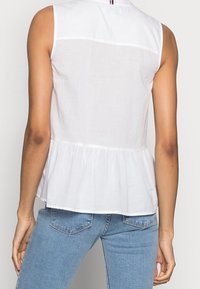 Top sans manches blanc avec un ourlet peplum, fabriqué en tissu léger. Coupe droite avec une encolure au dos et des détails de couture subtils.