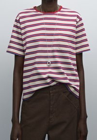 T-shirt a righe con linee rosse e blu su sfondo beige, dotata di collo rotondo e tasca; abbinata a pantaloni marroni e una collana con pendente.