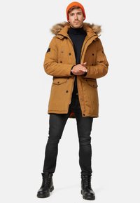 Parka marron avec capuche doublée de fourrure, boutons noirs et poches. Porté par-dessus un col roulé noir, associé à un jean foncé et des bottes noires.