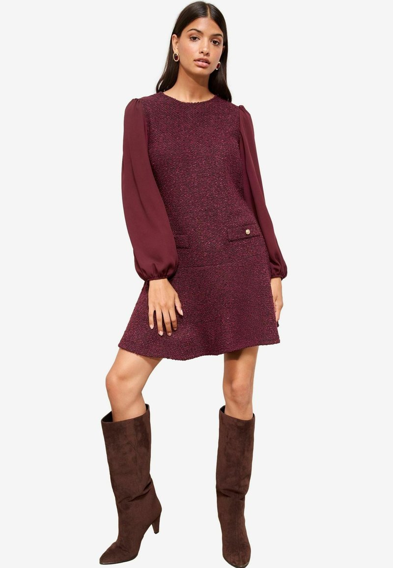 Rochie burgundy dintr-un material texturat, cu manșete lungi și translucide, două buzunare frontale și un buton decorativ aurie, asortată cu cizme înalte din suede maro.