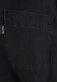 Tessuto di denim nero con una tasca posteriore cucita. Etichetta visibile vicino alla cucitura della tasca, che indica il marchio "Bershka".