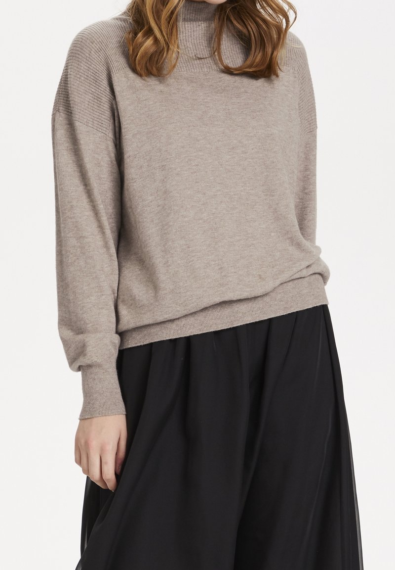 Ljus beige polotröja med ribbad textur vid axlar och ärmslut, med avslappnad passform och rak nederkant, parat med en svart kjol.