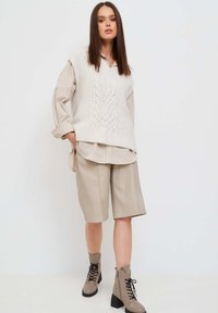 Gilet in maglia spessa con motivo a trecce, camicia color beige con colletto sotto, pantaloni corti beige su misura e stivali alla caviglia grigi con lacci neri.