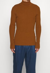 Maglione a collo alto color ruggine con coste verticali, vestibilità slim e maniche lunghe. Abbinato a pantaloni blu a gamba larga.