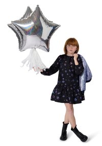 Mädchen in einem schwarzen Blumenkleid und Stiefeln hält drei silberne, sternförmige Ballons und eine silberne Jacke über der Schulter vor einem weißen Hintergrund.