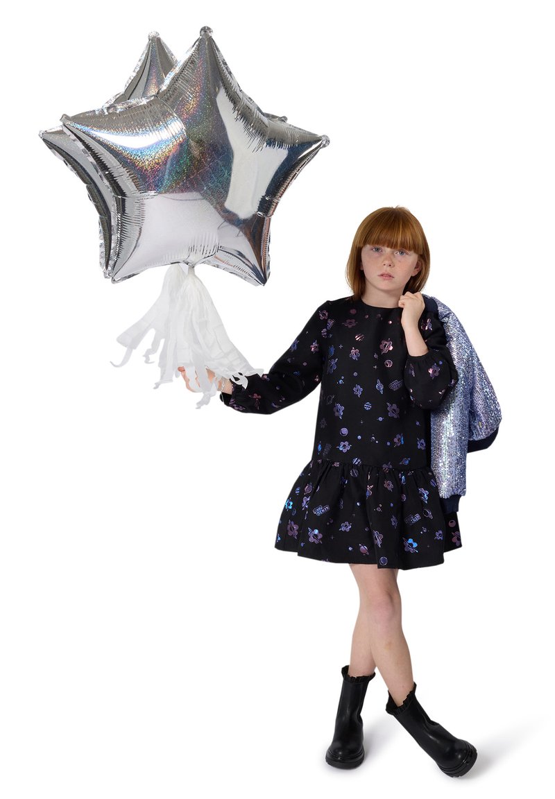 Mädchen in einem schwarzen Blumenkleid und Stiefeln hält drei silberne, sternförmige Ballons und eine silberne Jacke über der Schulter vor einem weißen Hintergrund.