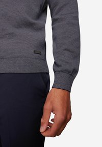 Dunkelblauer Strickpullover mit strukturierten Streifen, gerippten Bündchen und einem kleinen schwarzen Etikett nahe dem Saum. Nahaufnahme des Ärmels und der Hand.