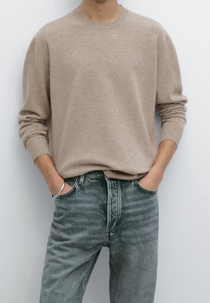 Personne portant un pull beige à manches longues rentré dans un jean gris délavé, les mains dans les poches, sur un fond uni.
