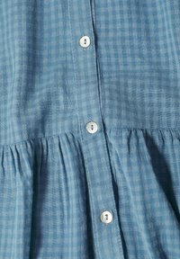 Chemise en tissu à carreaux bleus avec un devant à boutons, ornée de boutons ressemblant à des perles, avec un effet froncé à la taille et en matériau léger.
