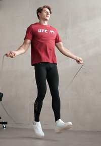 T-shirt rouge à manches courtes avec le logo "UFC", leggings noirs et baskets blanches. En train de sauter à la corde sur fond uni.