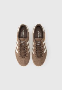 Καφέ σουέντ αθλητικά παπούτσια με τρεις λευκές ρίγες, στρογγυλή μύτη και υφασμένα κορδόνια. Η εσωτερική σόλα φέρει το λογότυπο της μάρκας για αναγνώριση.