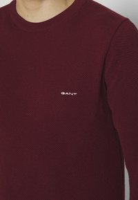 Bordeaux gebreide trui met een gestructureerde stof. Heeft een ronde halslijn en een klein wit "GANT" logo op de borst. Lange mouwen.