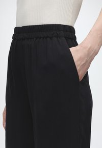 Άτομο που φοράει μαύρο παντελόνι με ελαστική μέση, με το δεξί χέρι στην τσέπη, συνδυασμένο με ένα ανοιχτόχρωμο ριπ μπλουζάκι.