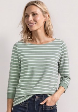 Lyshåret kvinde iført en grøn og hvid stribet langærmet skjorte, lidt stoppet ind i mørkeblå jeans, hånd i lommen, smilende og kigger mod højre.