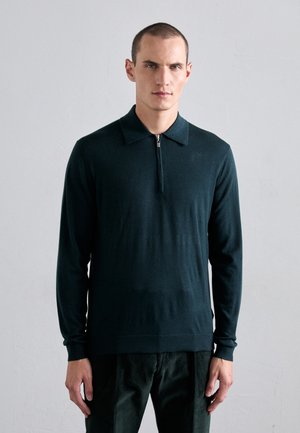 Hombre vistiendo una camisa polo de manga larga a rayas verde oscuro con cremallera y pantalones oscuros, de pie contra un fondo gris claro liso.