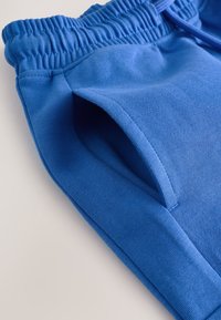 Modré bavlnené nohavice s elastickým pásom a bočným vreckom, zobrazené v detailnom záberu na svetlom pozadí.