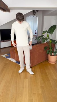 Camisa de manga larga color beige claro con un pequeño logo, combinada con pantalones crema y deportivas blancas. De pie sobre un suelo de madera en una habitación luminosa.