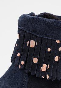 Botín de ante azul oscuro con detalles de flecos y acentos circulares metálicos en color oro rosado. Textura suave y borde superior redondeado.