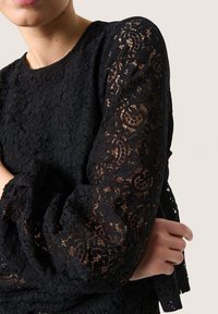 Top in pizzo nero con maniche lunghe e trasparenti decorate da motivi floreali. Il corpo è in pizzo nero solido, evidenziando texture e dettagli intricati.