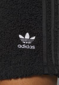 Svarta frottéshorts med vit Adidas-logotyp, tre svarta ränder längs sidorna och en texturerad, mjuk yta.