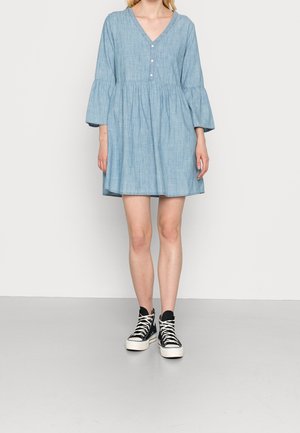 Femme portant une robe bleu clair à manches longues, col en V avec des boutons, et des baskets montantes noires avec des lacets et semelles blanches.