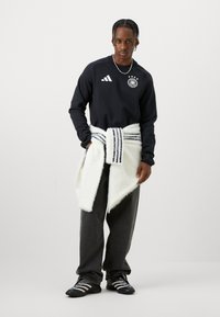 Mladic s pletenom kosom nosi crnu duksericu s bijelim emblemom, labave tamne hlače, crne tenisice i bijelu duksericu oko struka.