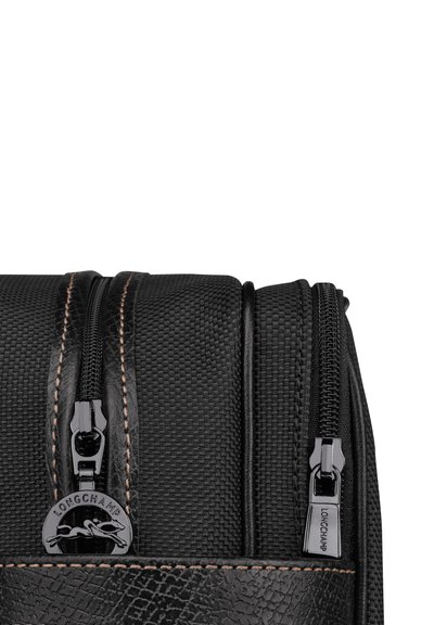 Schwarze, strukturierte Stofftasche mit schwarzen Metallreißverschlüssen, braunen Nähten und einem Logo-Element; rechteckige Form mit glatter Oberfläche.
