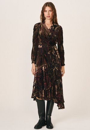 Femme portant une robe portefeuille sombre à motifs avec des manches longues et des bottes noires hautes jusqu’aux genoux, debout devant un fond clair uni.
