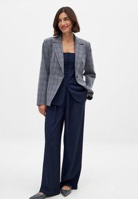 Grau karierter doppelt geknöpfter Blazer mit Revers, kombiniert mit einer marineblauen Hemdbluse und einer marineblauen weiten Hose. Flache graue Schuhe runden den Look ab.