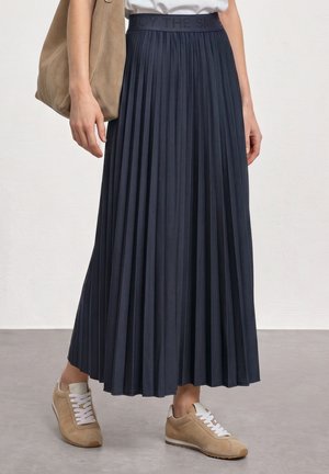 Femme portant une jupe midi plissée bleu marine avec des baskets beiges et tenant un grand sac à main beige à sa côté.