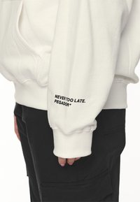 Vit hoodie med ribbad muddar, med svart text "ALDRIG FÖR SENT. PEGADOR*" på ärmen. Mjuk tyg, avslappnad passform, sidoficka.