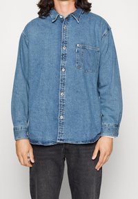 Persona che indossa una camicia in denim blu con bottoni e tasca sul petto, insieme a jeans neri, in piedi davanti a uno sfondo chiaro e uniforme.