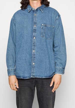 Persoon in een blauwe denim blouse met knoopsluiting en borstzak, gekleed in zwarte jeans, staande tegen een effen lichte achtergrond.