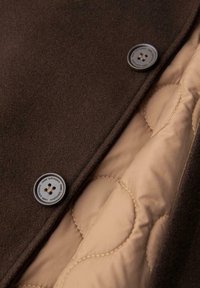 Ruskea villatakki, jossa on kaksi nappia ja vaalean beigen sävyinen sisäinen toppaus, sileä pinta ja kontrastiset tikkausompeleet.