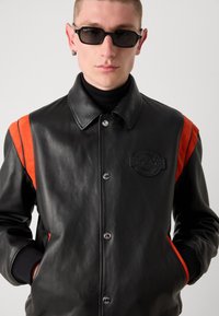 Sort læderjakke med orange suede-detaljer på skuldrene, trykknapper, ribbede manchetter og et struktureret emblem logo på brystet.