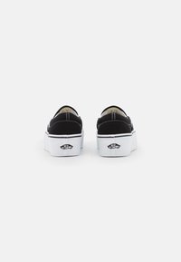 Black canvas slip-on shoes with a platform sole, featuring white rubber trim and logo detail on the back. Accents include contrasting stitching.