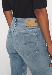 Jeans in denim azzurro chiaro con vita alta, cuciture a vista, due tasche posteriori e una etichetta in pelle beige che riporta le informazioni sul marchio nella cintura.
