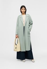 Γυναίκα που φοράει μακρύ ανοιχτό πράσινο παλτό, λευκό πλεκτό τοπ, φαρδιά σκούρα παντελόνια, λευκά παπούτσια, κρατώντας μια κρέμ τσάντα χειρός, στέκεται μπροστά σε απλό φόντο.