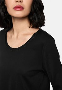 Schwarzes Langarmshirt mit rundem Ausschnitt, das einen subtilen goldenen Akzent entlang des Randes aufweist; aus einem glatten, weichen Stoff gefertigt.