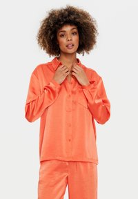 Camicia satinata color pesco, con colletto, maniche lunghe e tasca frontale. Indossata con pantaloni satinati abbinati.
