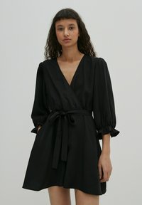 Zwarte wikkeljurk met een diepe V-hals, pofmouwen en een gestrikt taille. Gladde stof en een uitlopende rok die boven de knie eindigt.
