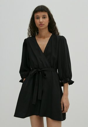Zwarte wikkeljurk met een diepe V-hals, pofmouwen en een gestrikt taille. Gladde stof en een uitlopende rok die boven de knie eindigt.
