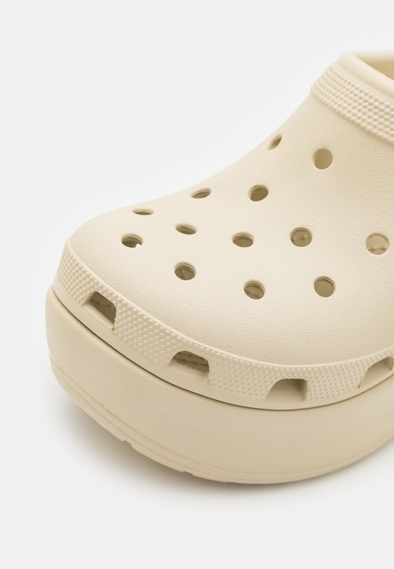 Zuecos beige hechos de material ligero con grandes agujeros de ventilación, una banda texturizada alrededor del borde superior y una suela elevada para mayor comodidad.
