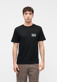 Νέος άνδρας που φοράει μαύρο t-shirt Billabong με μικρό λογότυπο τοπίου στο στήθος και καφέ παντελόνι, στέκεται μπροστά σε λευκό φόντο.
